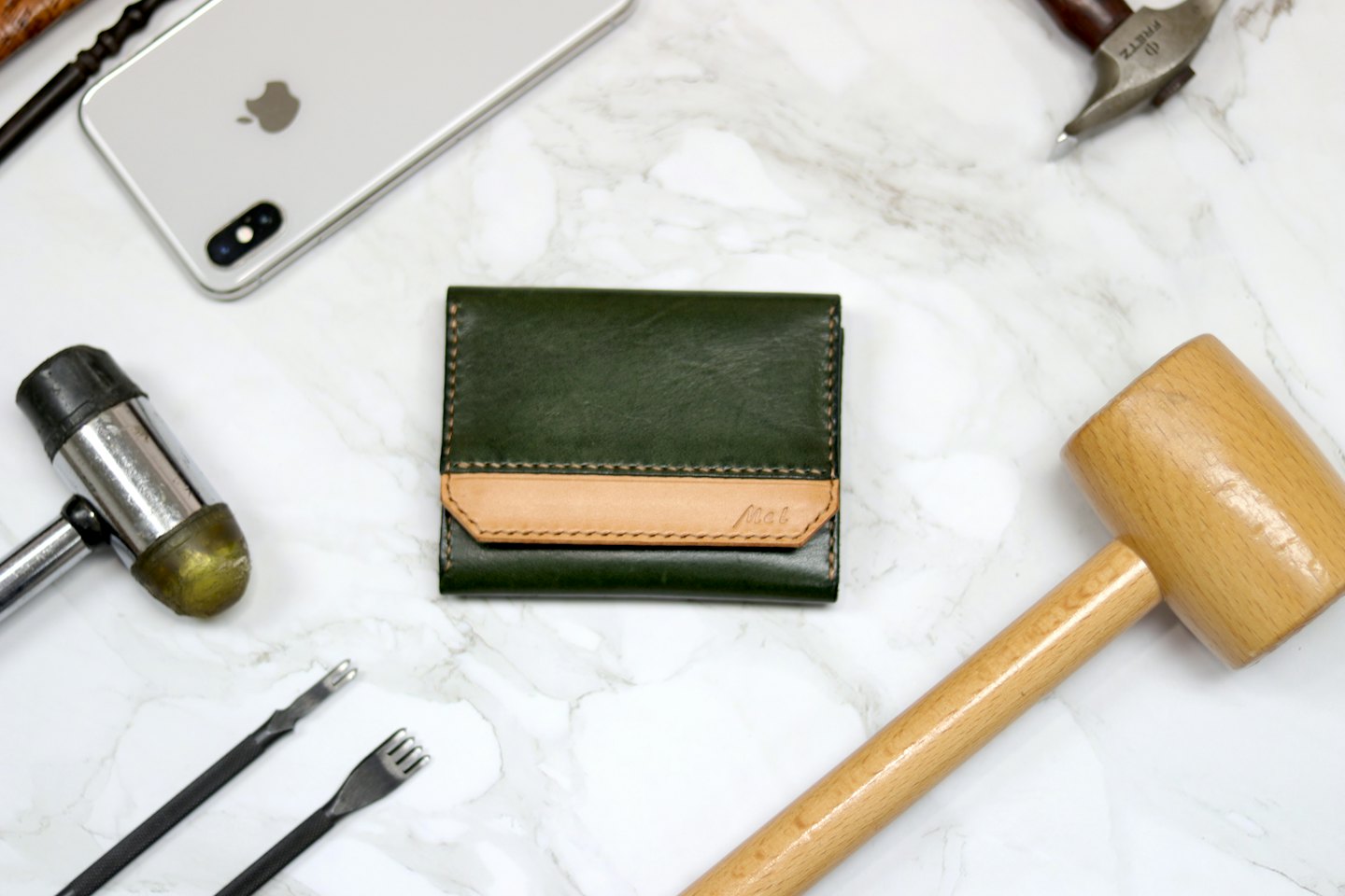 A handmade leather wallet surrounded by various leatherworking tools on marble.